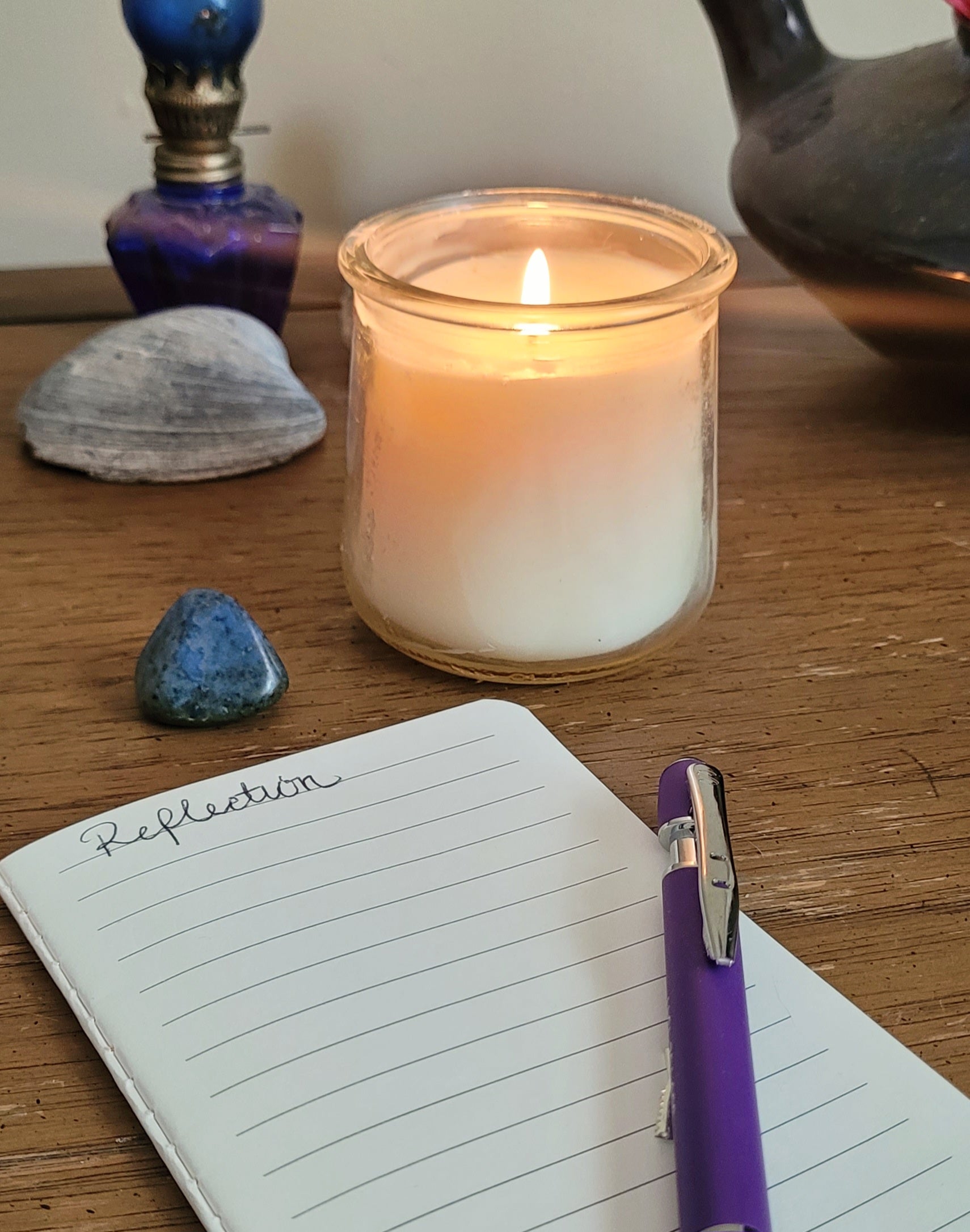 Candle in a glass jar, notebook with 'Reflection' title, and purple pen on a wooden surface.