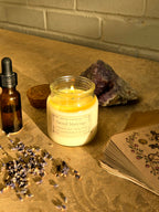 A lit candle in a clear glass jar sits on a stone surface with a small amber dropper bottle, dried lavender, a crystal, and a stack of illustrated cards against a brick wall background.