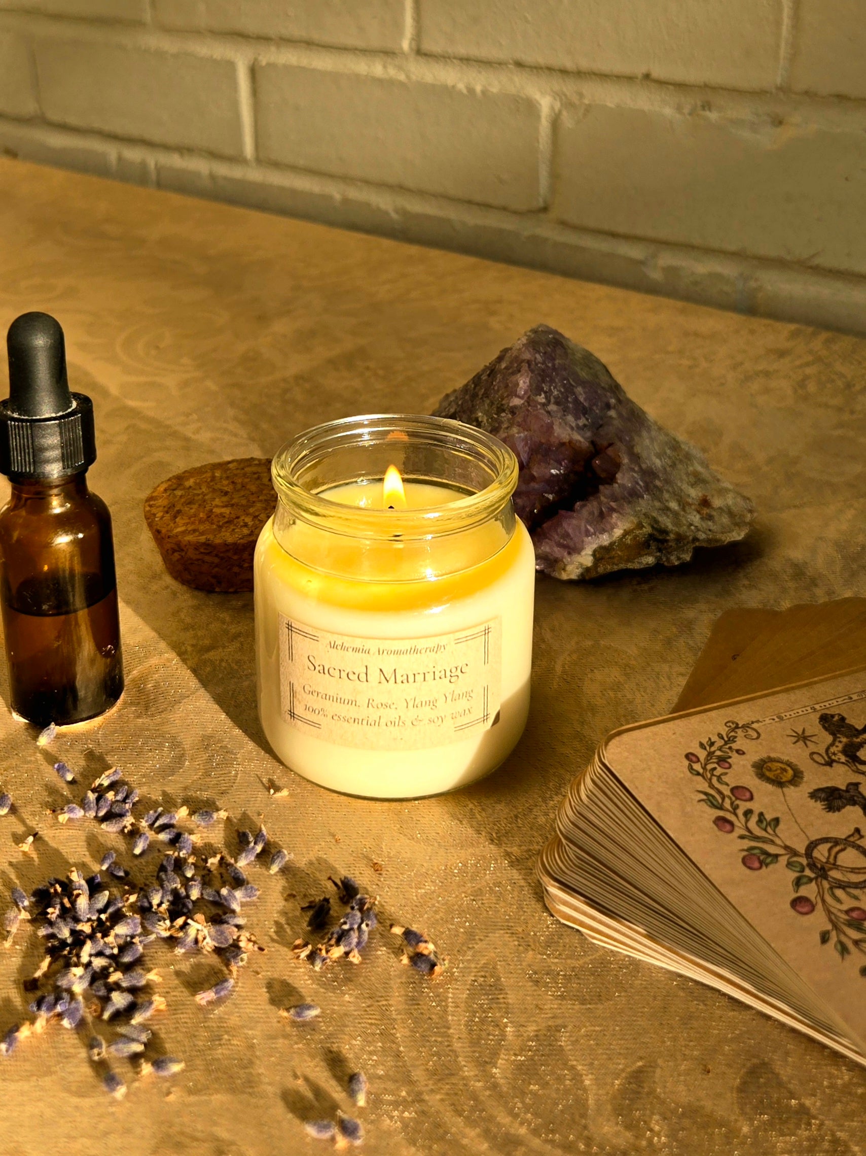 A lit candle in a clear glass jar sits on a stone surface with a small amber dropper bottle, dried lavender, a crystal, and a stack of illustrated cards against a brick wall background.