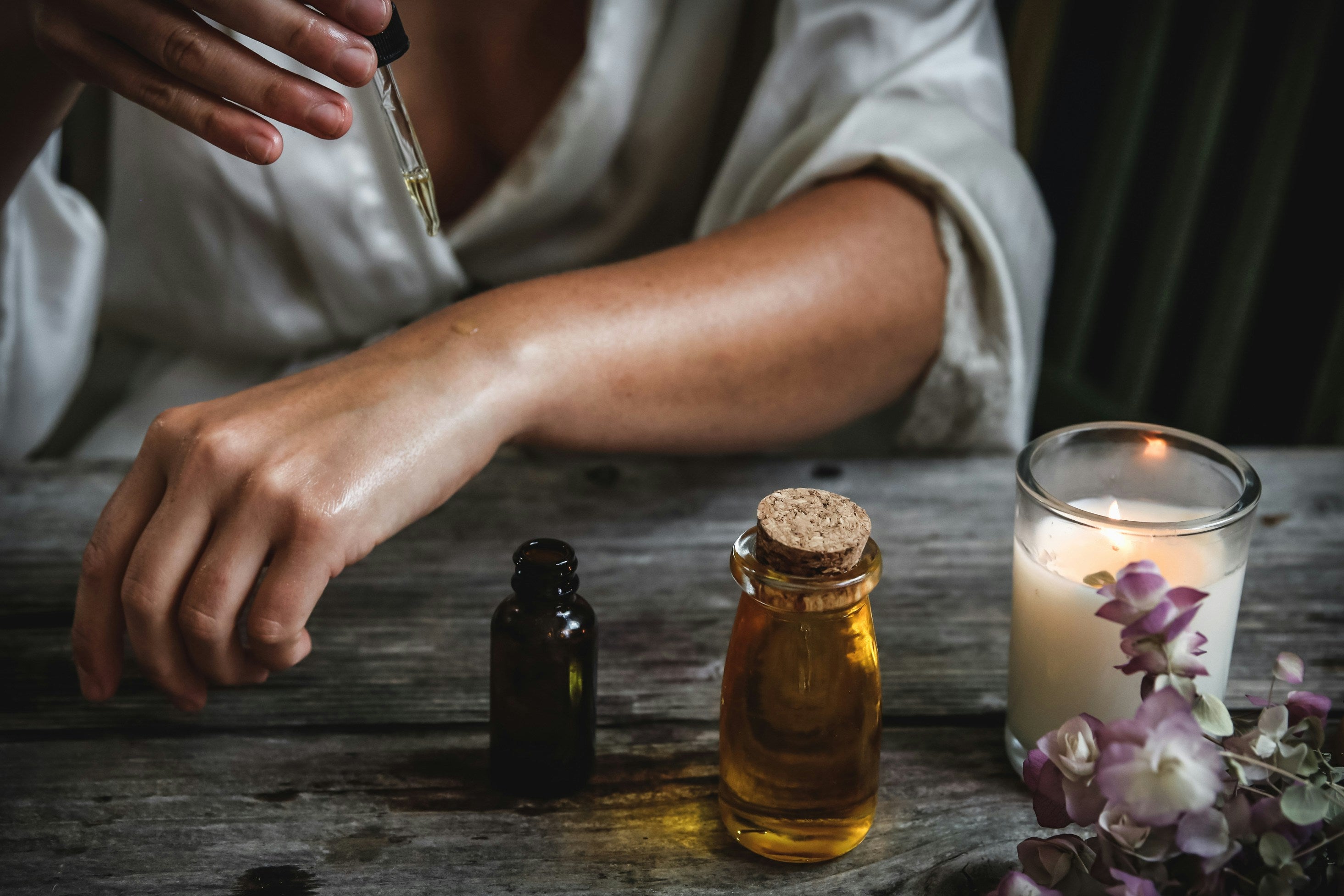 Person holding a dropper over bottles on a wooden surface with a lit candle and flowers.