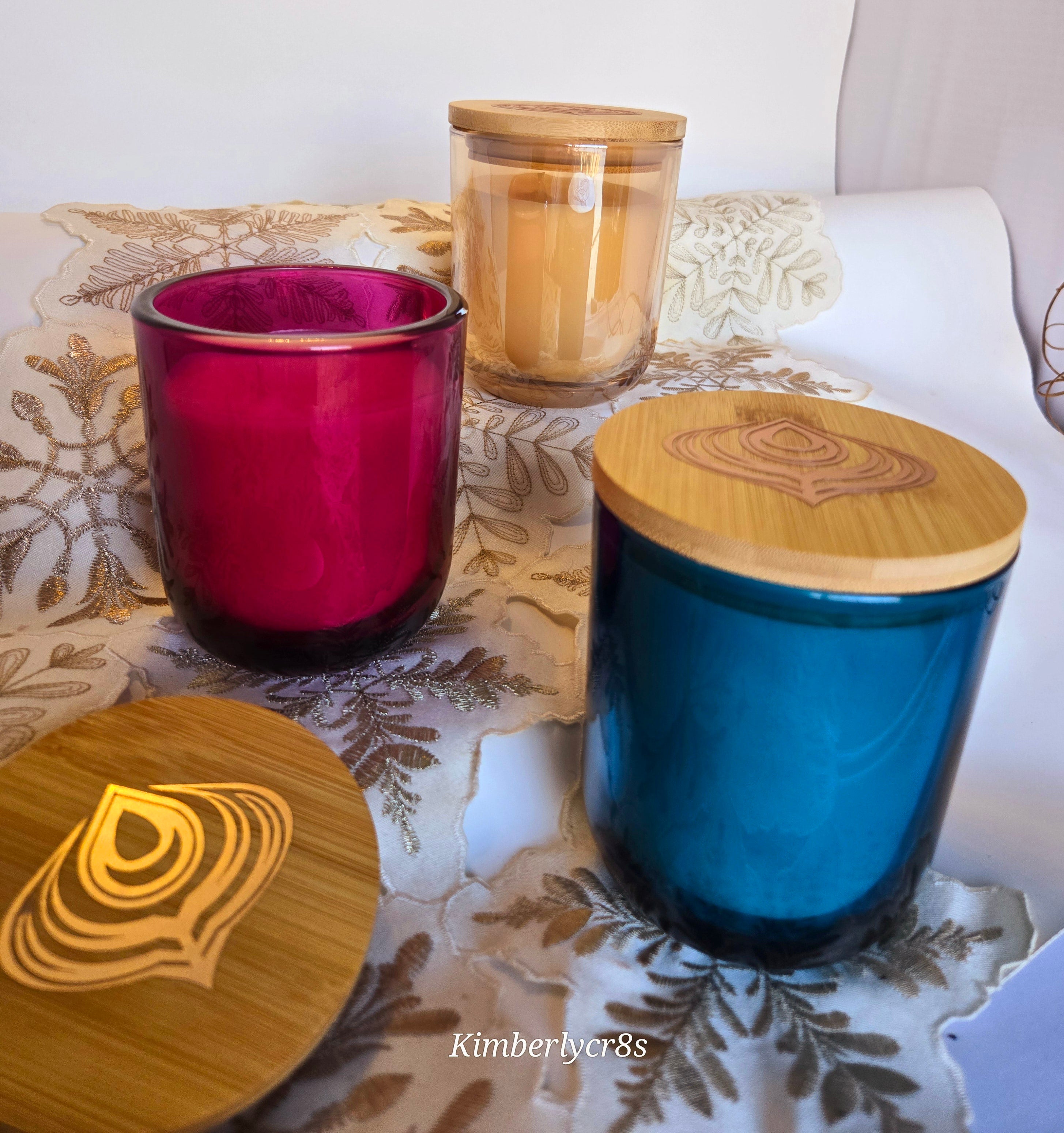 Three candles with colorful glass jars, each with a bamboo lid, placed on a surface with a patterned fabric.