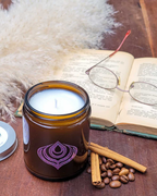 Candle in a brown jar with a purple logo on a wooden surface with an open book, glasses, and cinnamon sticks.