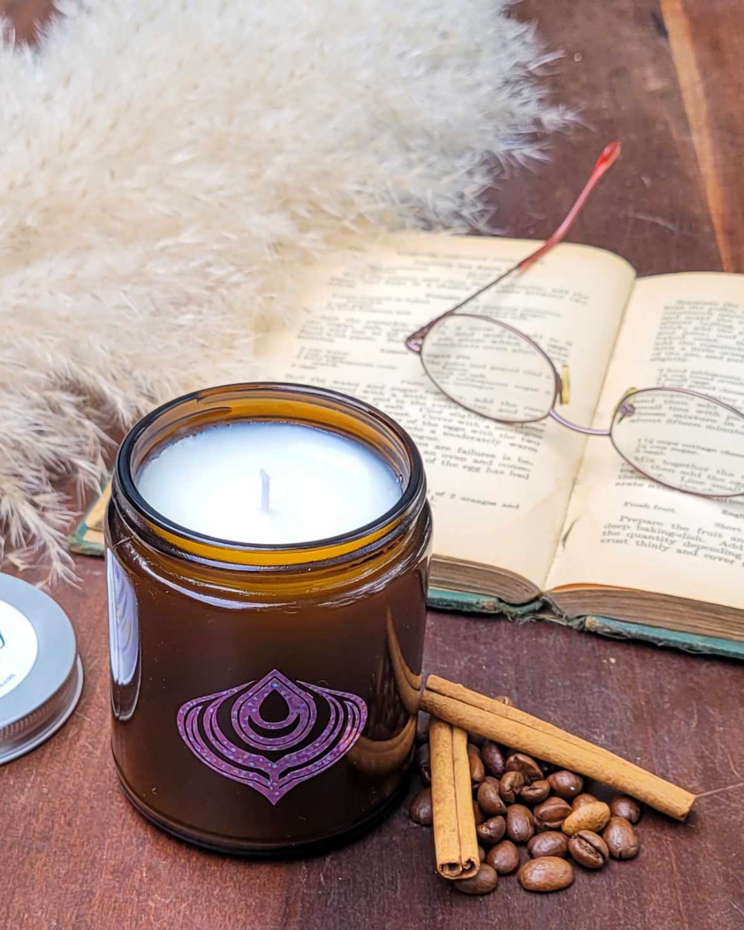 Candle in a brown jar with a purple logo on a wooden surface with an open book, glasses, and cinnamon sticks.