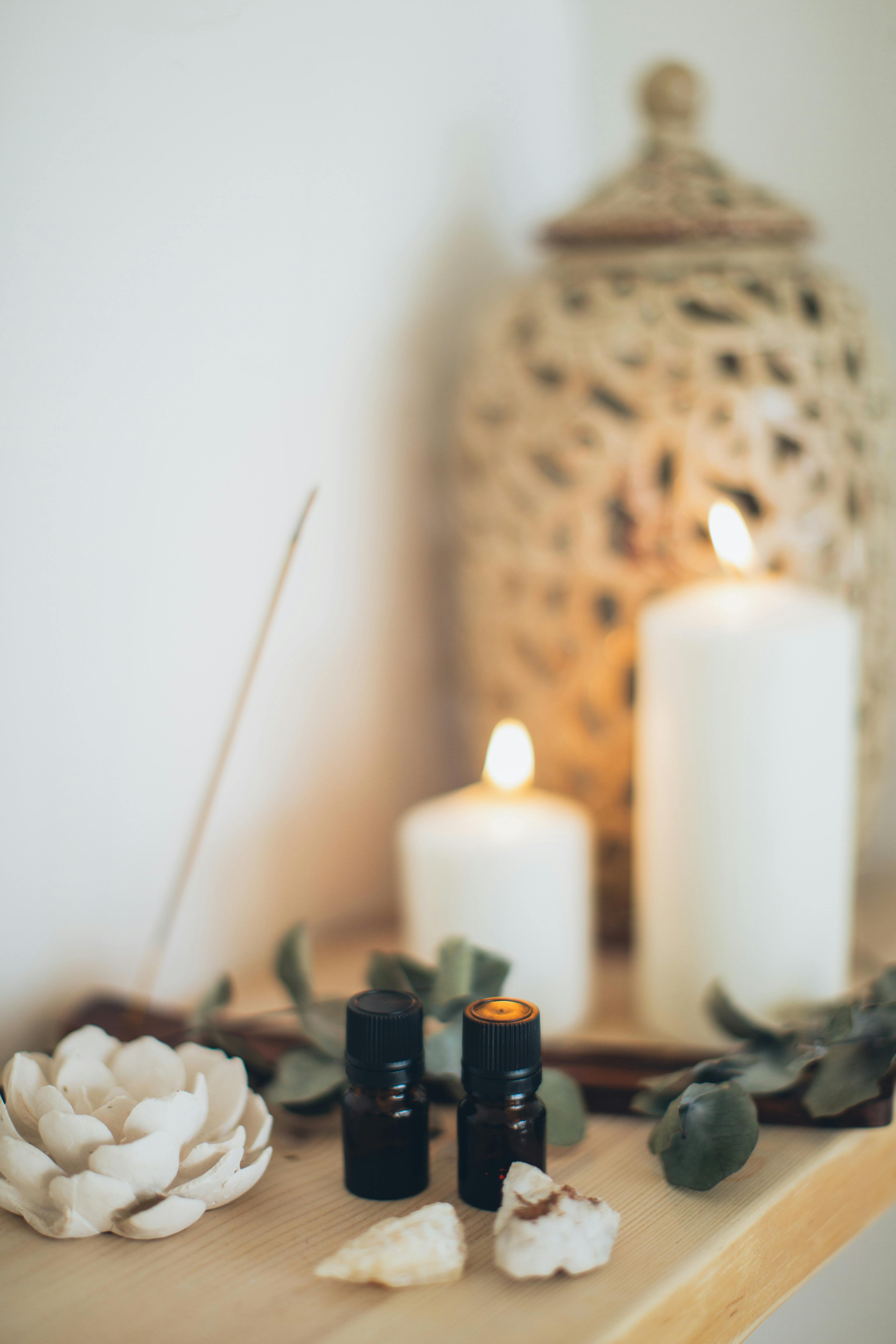 Two lit candles, two small black bottles, a white flower, and a woven jar on a wooden surface.