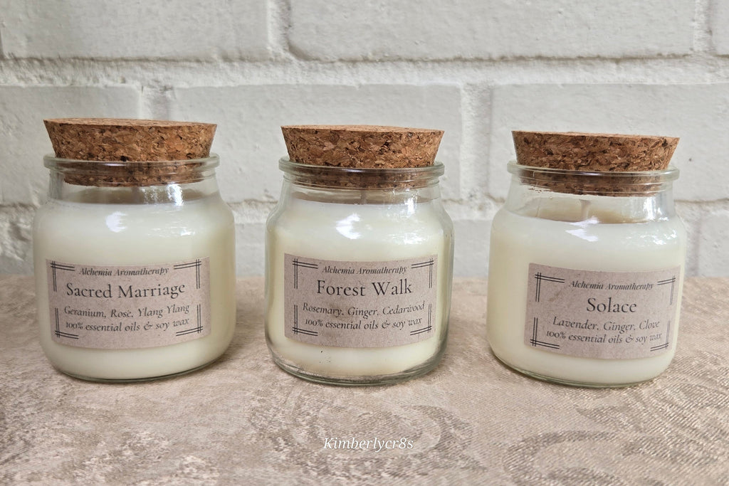 Three glass jar candles with cork lids are lined up on a textured surface against a white brick wall. The labels read 'Sacred Marriage', 'Forest Walk', and 'Solace'.