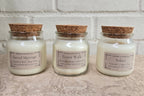 Three glass jar candles with cork lids are lined up on a textured surface against a white brick wall. The labels read 'Sacred Marriage', 'Forest Walk', and 'Solace'.