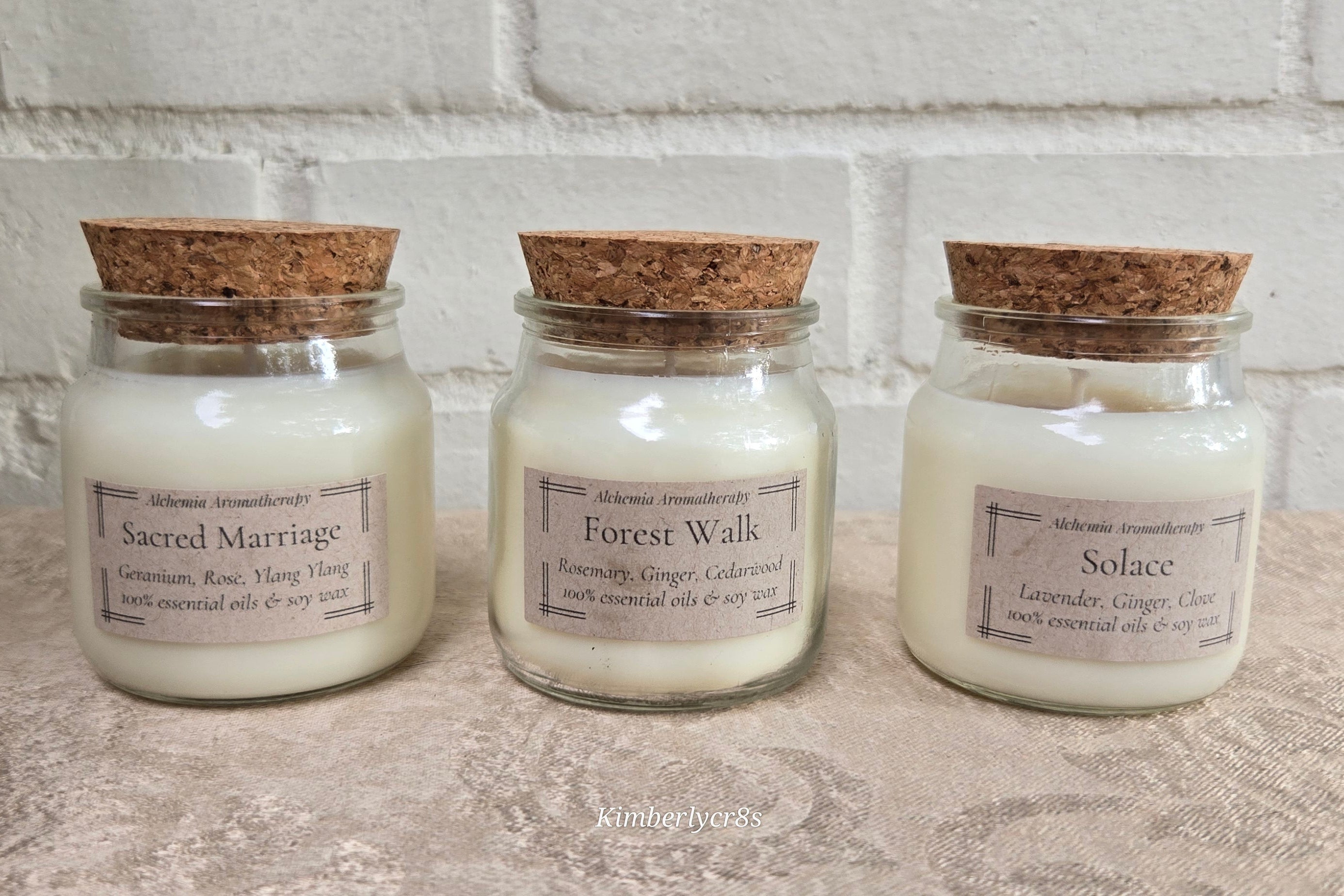 Three glass jar candles with cork lids are lined up on a textured surface against a white brick wall. The labels read 'Sacred Marriage', 'Forest Walk', and 'Solace'.