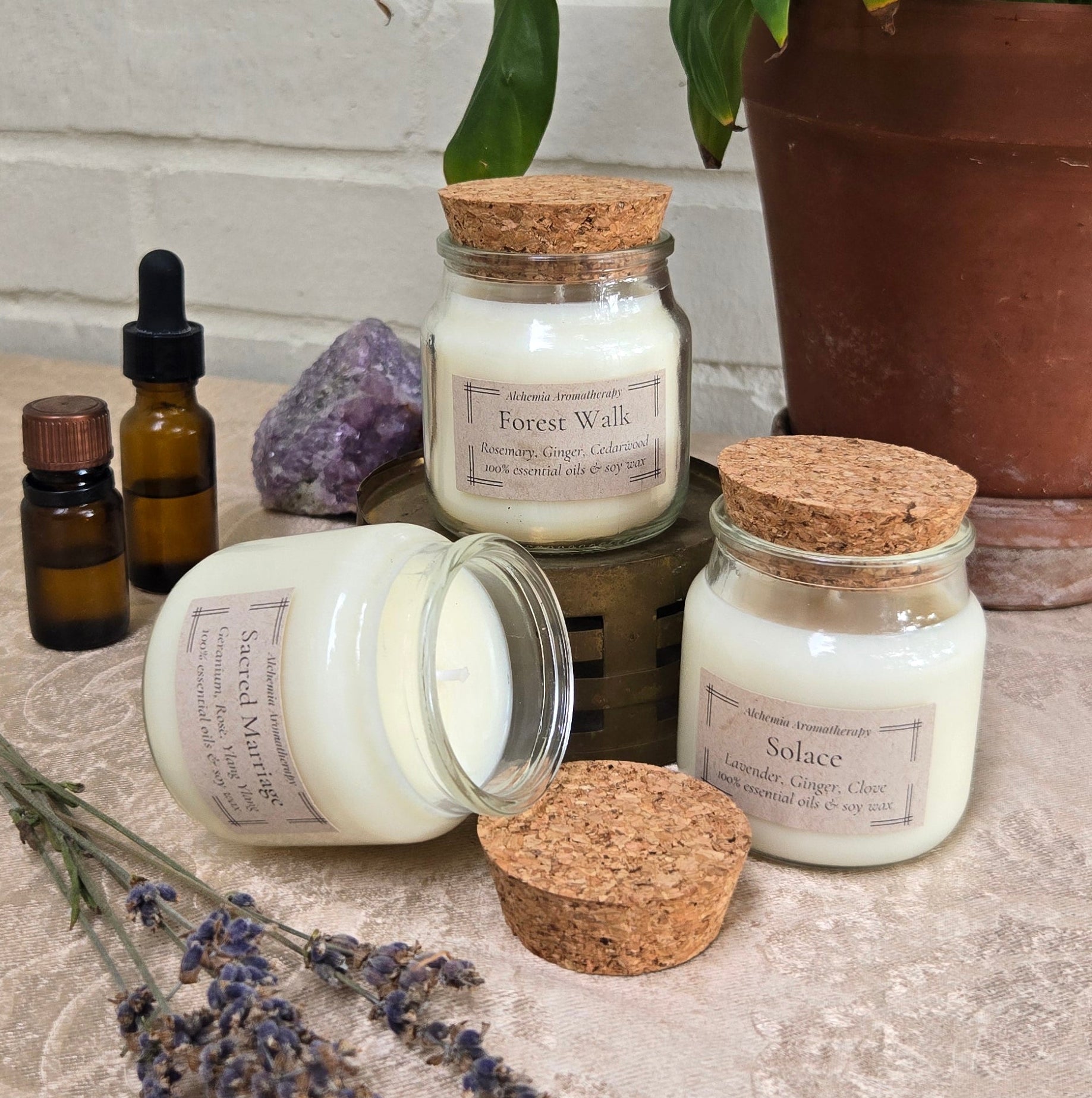 Three white wax candles in glass jars with cork lids are arranged on a textured beige surface with a potted green plant, purple crystals, amber dropper bottles, and dried lavender sprigs. One candle jar is tipped over, showing the white wax.